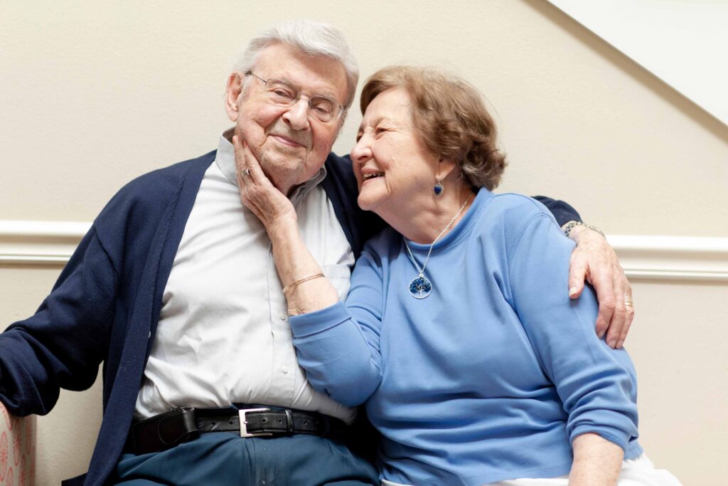 A senior couple in sweats of different shades of blue sit together. The man on the left is smiling at the camera. The woman on the right has her hand on his cheek as she smiles over at him.