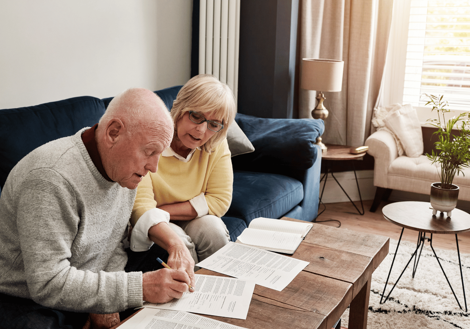 Elderly couple sitting on a sofa at home, reviewing papers and a notebook on a wooden coffee table.