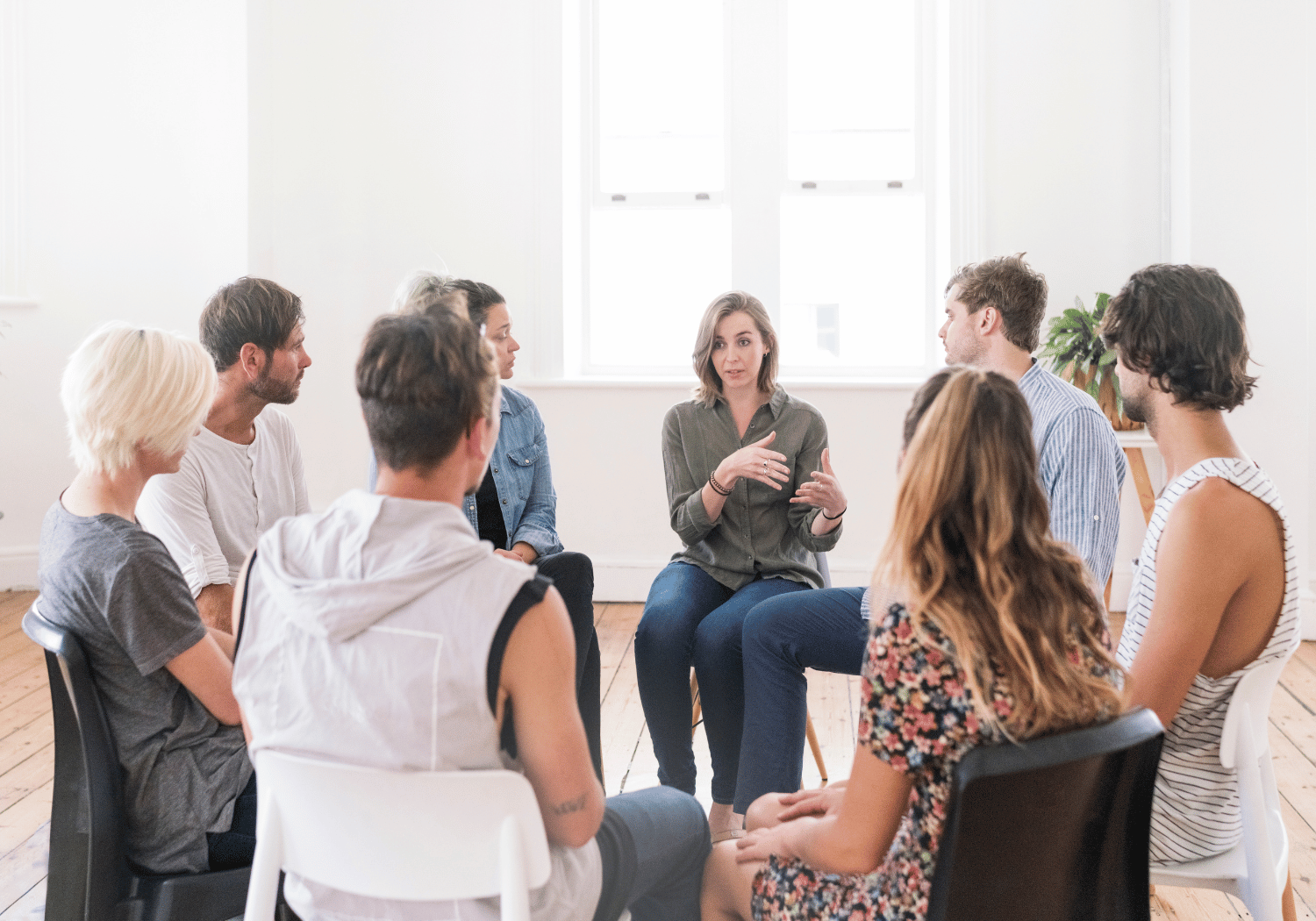 Group of adults sitting in a circle, a woman in the center speaks while the others listen in a bright, minimalist room.