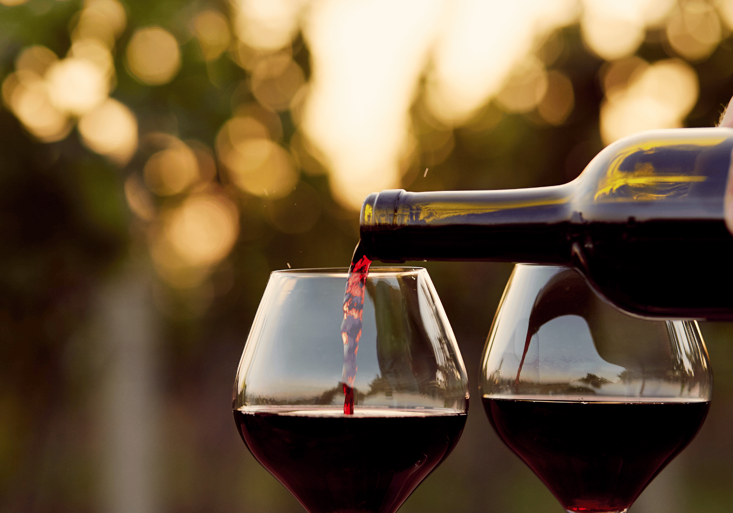Red wine being poured from a bottle into two stemmed glasses outdoors at sunset.